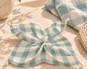 May include: A hair scrunchie featuring a light blue and white gingham pattern with small embroidered blue fish motifs. The fabric is gathered at the center and finished with four white pearl-like beads attached to the corners of the fabric ends.
