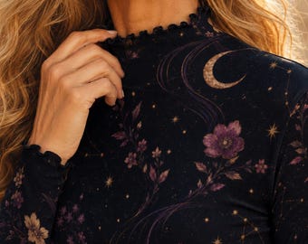 May include: A long-sleeved mock neck top featuring a dark navy background with a celestial and floral print. The pattern includes purple flowers, golden stars, and a crescent moon. The garment has a ruffled lettuce-edge trim at the neck and cuffs.