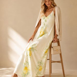 A woman in a lemon-print maxi dress and a cream cardigan sits on a stool with a small cream purse.