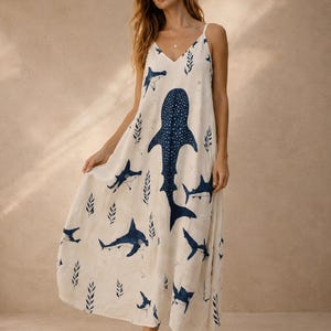 Puede incluir: Un vestido largo sin mangas de color crema con escote en V y tirantes finos. La tela está decorada con un patrón repetitivo de tiburones y tiburones ballena en azul oscuro, intercalados con pequeños motivos de hojas en azul oscuro.