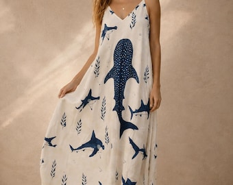 May include: A sleeveless, floor-length cream-colored dress featuring a V-neckline and thin spaghetti straps. The fabric is decorated with a repeating pattern of dark blue sharks and whale sharks, interspersed with small dark blue leaf motifs.
