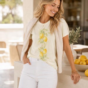 Op de afbeelding: Een lichtgeel t-shirt met korte mouwen en een verticale botanische illustratie van felgele citroenen met groene bladeren en witte bloesems in het midden, gecombineerd met een witte spijkerbroek en een beige gebreide trui over de schouders.