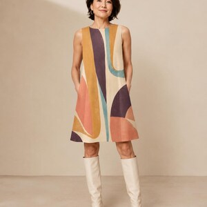 May include: A sleeveless, knee-length shift dress featuring a cream base with large, abstract, curved shapes in shades of mustard yellow, deep purple, and muted coral. The dress has a round neckline and side pockets, paired with cream knee-high boots.