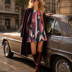 May include: A sleeveless mini dress featuring a geometric pattern in shades of pink, navy blue, and cream, worn with a dark purple overcoat and dark red knee-high boots. The dress has a round neckline and a flared silhouette, showcasing abstract shapes.