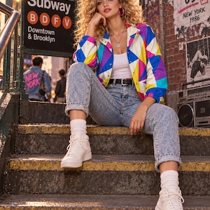 Puede incluir: Una chaqueta cortavientos con estampado geom&eacute;trico multicolor y cierre de botones a presi&oacute;n, sobre una camiseta sin mangas blanca, combinada con vaqueros azules de lavado &aacute;cido, calcetines blancos y zapatillas altas blancas, con collar y aros rosas.