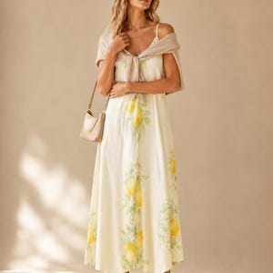 A woman in a lemon-print maxi dress has a beige knit sweater draped over her shoulders and carries a small beige purse.