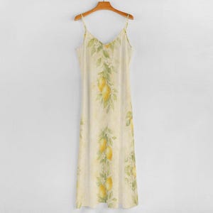 A light yellow spaghetti strap maxi dress with a lemon and leaf print hangs on a wooden hanger against a gray wall.