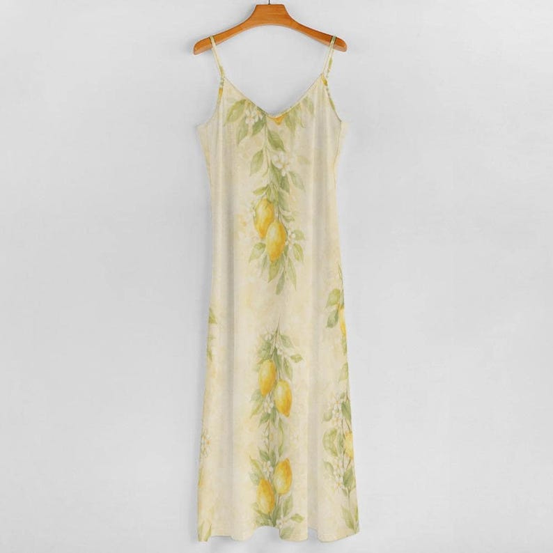 A light yellow spaghetti strap maxi dress with a lemon and leaf print hangs on a wooden hanger against a gray wall.