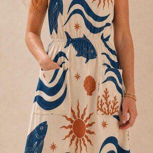 May include: A sleeveless cream-colored dress featuring a repeating print of blue fish, blue waves, orange suns, orange seashells, and orange coral. The dress has a V-neckline, an elasticized waist, and two large patch pockets on the front skirt.