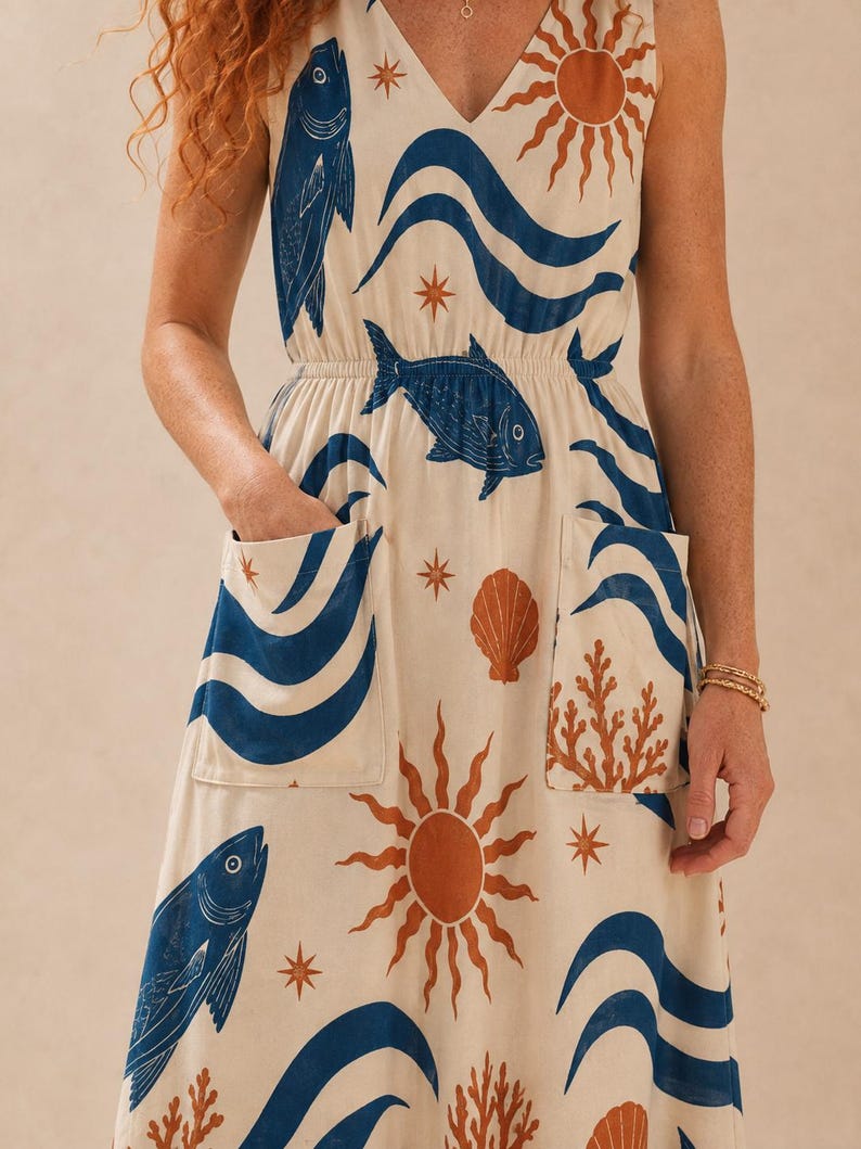 May include: A sleeveless cream-colored dress featuring a repeating print of blue fish, blue waves, orange suns, orange seashells, and orange coral. The dress has a V-neckline, an elasticized waist, and two large patch pockets on the front skirt.