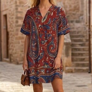 K&ouml;nnte beinhalten: Kurz&auml;rmeliges V-Ausschnitt-Kleid mit Paisley-Muster in Rot-, Blau- und Goldt&ouml;nen. Das Kleid hat einen blauen Saum an den &Auml;rmeln und am Saum. Eine kleine braune Handtasche wird in der Hand gehalten.