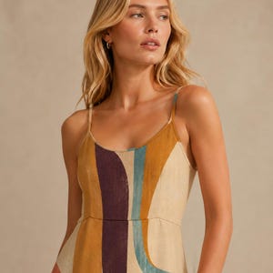 May include: A sleeveless dress featuring a scoop neckline and thin spaghetti straps. The garment is decorated with a bold, abstract pattern of vertical and curved bands in shades of mustard yellow, deep purple, and muted teal on a beige base.