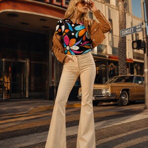 May include: A short-sleeved t-shirt with a black background and a colorful floral pattern in orange, blue, pink, and white. The shirt is paired with a tan cropped jacket and cream-colored flared trousers. The outfit includes brown platform sandals.