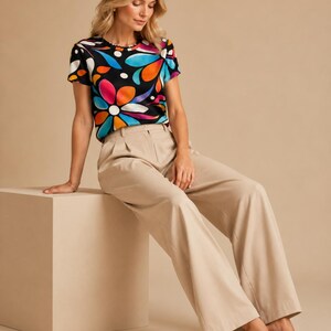 May include: A short-sleeved crew neck t-shirt featuring a vibrant, multicolored floral and abstract pattern on a black background, paired with high-waisted, wide-leg beige trousers that have a pleated front and a straight-leg silhouette.