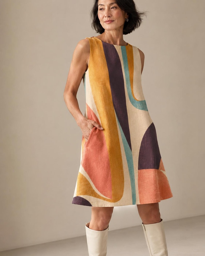 May include: A sleeveless knee-length shift dress featuring a bold abstract pattern with large, flowing shapes in shades of mustard yellow, deep purple, light blue, and coral orange on a cream-coloured background, complete with side pockets.