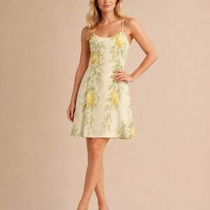 May include: A sleeveless mini dress in a pale yellow hue featuring a repeating pattern of bright yellow lemons and green leafy branches. The dress has a scoop neckline, thin spaghetti straps, and a fitted bodice that flares into a skirt.