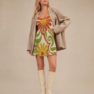 May include: A sleeveless mini dress featuring a large-scale floral and swirling abstract pattern in shades of orange, yellow, olive green, and cream, worn under an oversized beige blazer with long sleeves and white knee-high boots.