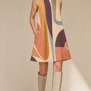 May include: A sleeveless A-line dress featuring a cream base with large, abstract curved shapes in shades of mustard yellow, deep purple, terracotta orange, and muted teal. The dress has a round neckline and side pockets, paired with beige boots.