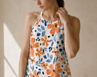 May include: A sleeveless white halter top featuring a watercolor-style floral print with large orange flowers and dark blue leaves. The garment has a high neckline with thin straps that extend over the shoulders, creating a clean and simple silhouette.