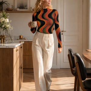 May include: A long-sleeve mock neck top featuring a wavy, multi-colored pattern in shades of orange, green, and black, paired with high-waisted, wide-leg cream trousers that feature a pleated front and a clean, tailored silhouette.