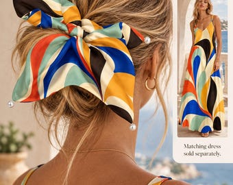 May include: A hair accessory featuring a multi-colored abstract print in shades of blue, orange, yellow, black, and cream. The fabric is tied into a bow shape and accented with small white pearl-like beads at the ends. Text reads: Designed in the same print as our matching dress. Matching dress sold separately.
