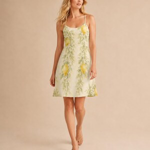 May include: A sleeveless knee-length dress featuring a light cream background with a repeating vertical pattern of yellow lemons and green leafy vines. The garment has a scoop neckline and thin spaghetti straps, with a straight-cut hemline.