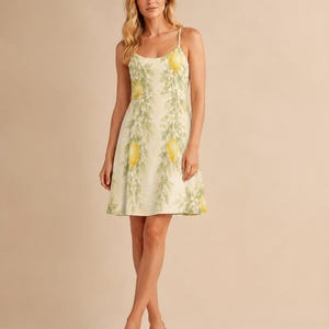 May include: A sleeveless knee-length dress featuring a light cream background with a repeating pattern of yellow lemons and green leafy branches. The garment has thin spaghetti straps, a scoop neckline, and a flared skirt silhouette.
