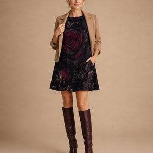 Puede incluir: Un vestido corto negro sin mangas con un estampado de una gran rosa de color rojo oscuro, usado bajo un blazer color canela con mangas tres cuartos. El conjunto se combina con botas altas de cuero color burdeos oscuro con punta fina.