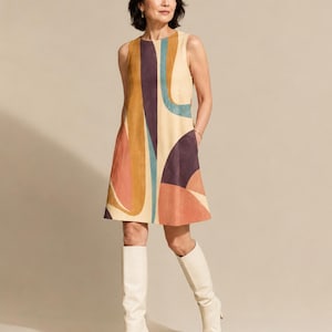 May include: A sleeveless knee-length shift dress featuring a cream base with large abstract shapes in shades of mustard yellow, deep purple, teal, and terracotta. The dress has a round neckline and side pockets, paired with cream knee-high boots.