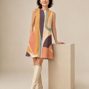 May include: A sleeveless A-line dress featuring a bold abstract pattern with large curved shapes in shades of muted orange, deep purple, teal, and cream. The dress has a round neckline and a straight hemline that falls above the knee.