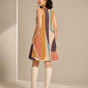 May include: A sleeveless, knee-length shift dress featuring a bold, abstract pattern of curved shapes in shades of mustard yellow, terracotta, deep purple, and teal on a cream background, complete with side pockets and paired with cream knee-high boots.