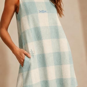 May include: A sleeveless, light blue and white checkered dress with a small fish emblem near the neckline and a leaf emblem near the pocket. The dress has a relaxed fit and a scoop neckline.