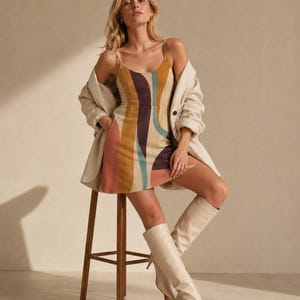 May include: A sleeveless mini dress featuring a scoop neckline and a multi-colored abstract pattern in shades of mustard yellow, deep purple, and teal on a beige background, paired with an oversized cream-colored blazer and cream knee-high boots.