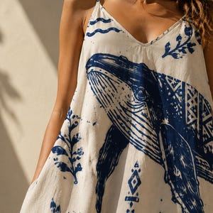 May include: White sundress with a blue whale illustration. The dress features a V-neck and thin straps. Decorative blue patterns of waves, leaves, and geometric shapes are printed on the fabric. The dress is a casual summer garment.