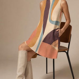 May include: A sleeveless mini dress featuring a bold, abstract geometric pattern in shades of terracotta, deep purple, and muted blue on a cream-coloured base. The dress has a round neckline, a straight silhouette, and visible side pockets.