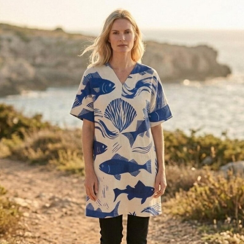 Puede incluir: Vestido color crema de manga corta y cuello en V, con un estampado de peces y conchas marinas azules. El vestido se lleva con pantalones negros. El fondo muestra un paisaje costero con una masa de agua y terreno rocoso.
