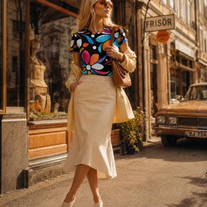 May include: A short-sleeved t-shirt featuring a vibrant, multi-colored floral pattern on a black background, paired with a high-waisted, cream-colored midi skirt with a flared hem, a light beige jacket, a brown shoulder bag, and open-toe sandals.