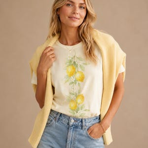Op de afbeelding: Een cr&egrave;mekleurig T-shirt met korte mouwen en een verticale botanische print van gele citroenen en groene bladeren in het midden, gecombineerd met een lichtgeel gebreide trui over de schouders en een lichtblauwe spijkerbroek.