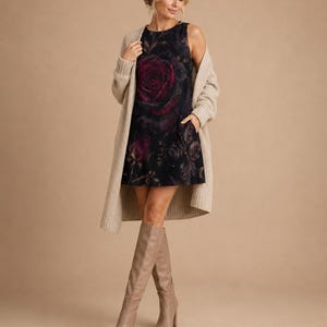 Puede incluir: Un vestido corto sin mangas de color negro con un estampado de rosas grandes en rojo oscuro, combinado con un c&aacute;rdigan largo de punto acanalado en color beige y botas altas de cuero beige. El vestido tiene cuello redondo y bolsillos laterales.
