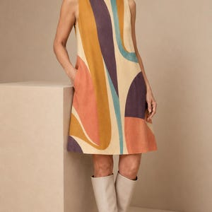 May include: A sleeveless shift dress featuring a cream base with large, abstract, curved shapes in shades of mustard yellow, deep purple, terracotta orange, and teal blue. The dress has a round neckline and side pockets, paired with knee-high boots.