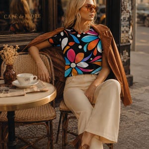 May include: A short-sleeved t-shirt featuring a black background with a vibrant, multi-colored floral and petal pattern in shades of blue, orange, pink, and white, paired with beige trousers and a brown jacket draped over the shoulders.