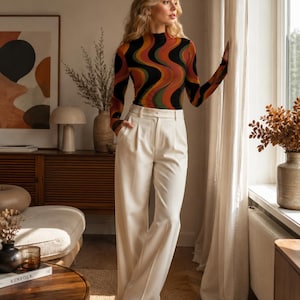 May include: A long-sleeve mock neck top featuring a wavy, multi-colored pattern in shades of black, orange, yellow, and green, paired with high-waisted, wide-leg cream trousers that feature a front pleat and a structured waistband.