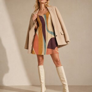 May include: A sleeveless mini dress featuring a multi-colored abstract pattern in shades of mustard, deep purple, teal, and terracotta. The dress is paired with a beige blazer draped over the shoulders and cream-colored knee-high heeled boots.