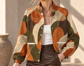 Puede incluir: Una chaqueta de manga larga con un patrón abstracto multicolor en tonos naranja quemado, verde oliva y crema. La chaqueta tiene cuello alto, botones de presión plateados y dos bolsillos delanteros con ribete blanco, usada sobre una camiseta blanca.