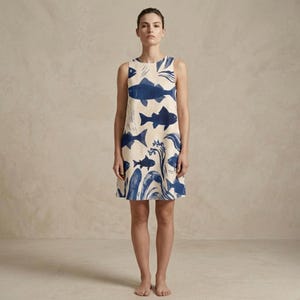 A woman stands barefoot, facing forward, wearing a sleeveless A-line dress with a blue fish and wave pattern on a cream background.
