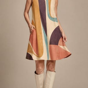 May include: A sleeveless A-line dress featuring a bold abstract pattern in shades of mustard yellow, deep purple, muted teal, and terracotta orange on a cream background, with a round neckline and side pockets, paired with cream knee-high boots.