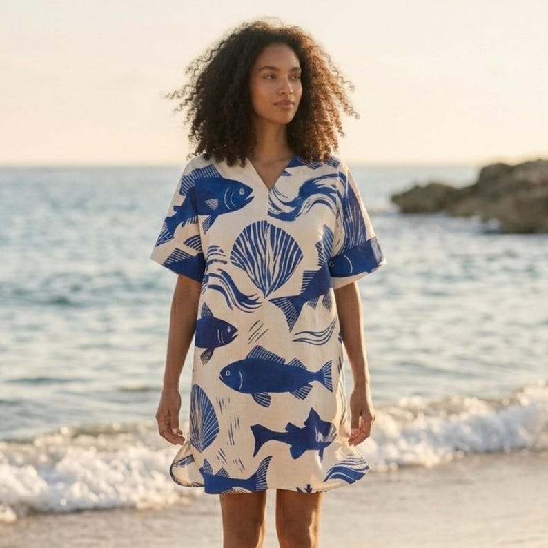 Puede incluir: Vestido de manga corta con cuello en V, con fondo crema y estampado de peces y conchas marinas azules. El vestido tiene un dobladillo con volantes y se lleva en una playa con el oc&eacute;ano al fondo.