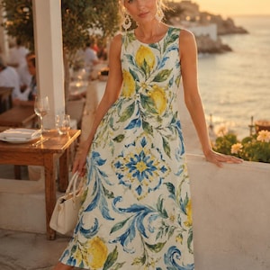 May include: A sleeveless midi dress featuring a white base with a vibrant print of yellow lemons, green leaves, and blue decorative scrollwork patterns. The dress has a round neckline and a fitted bodice that flows into a relaxed skirt silhouette.