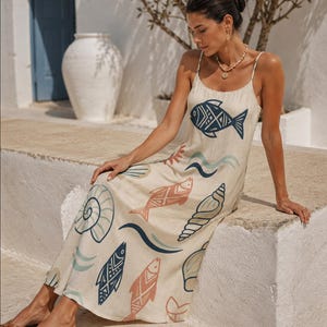 May include: A beige sleeveless midi dress featuring thin spaghetti straps and a repeating pattern of stylized fish, seashells, and wave motifs in shades of navy blue, light blue, and terracotta orange printed across the fabric.