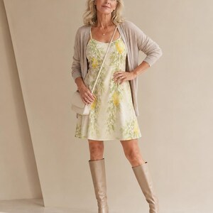 May include: A sleeveless mini dress featuring a yellow and green floral print, worn with a beige open-front cardigan, a small beige crossbody bag, and beige knee-high boots. The dress has a scoop neckline and a fitted bodice with a flared skirt.